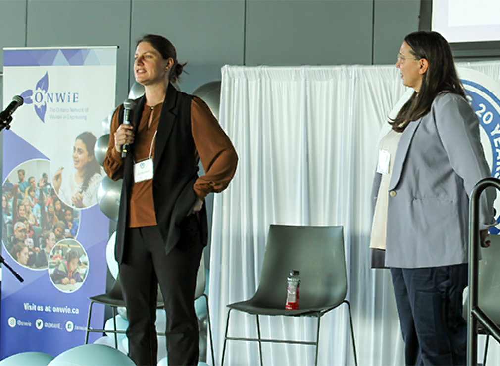 Kim Bouffard and Rebecca White stand on a stage speaking at a conference; one holds a microphone while the other listens. A banner for ONWiE is visible in the background.